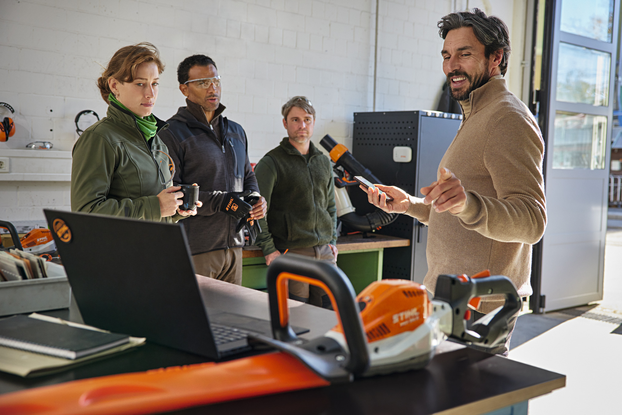 Fire personer står i et værksted, mens en person forklarer STIHL connected-appen på en smartphone