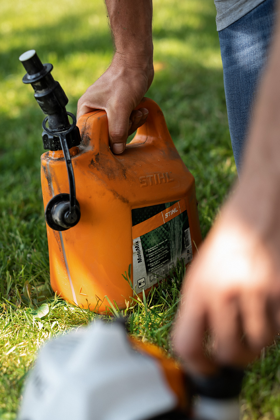 En snavset dunk med STIHL MotoMix-brændstofblanding på græs med en hånd, der åbner brændstofdækslet på et værktøj i forgrunden